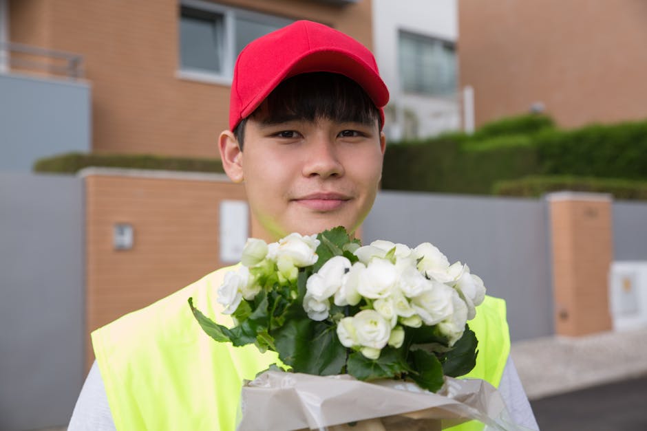 A young delivery person wearing a red cap and a high-visibility yellow vest is holding a bouquet of fresh white flowers wrapped in brown paper. The bouquet consists of small, tightly clustered blossom