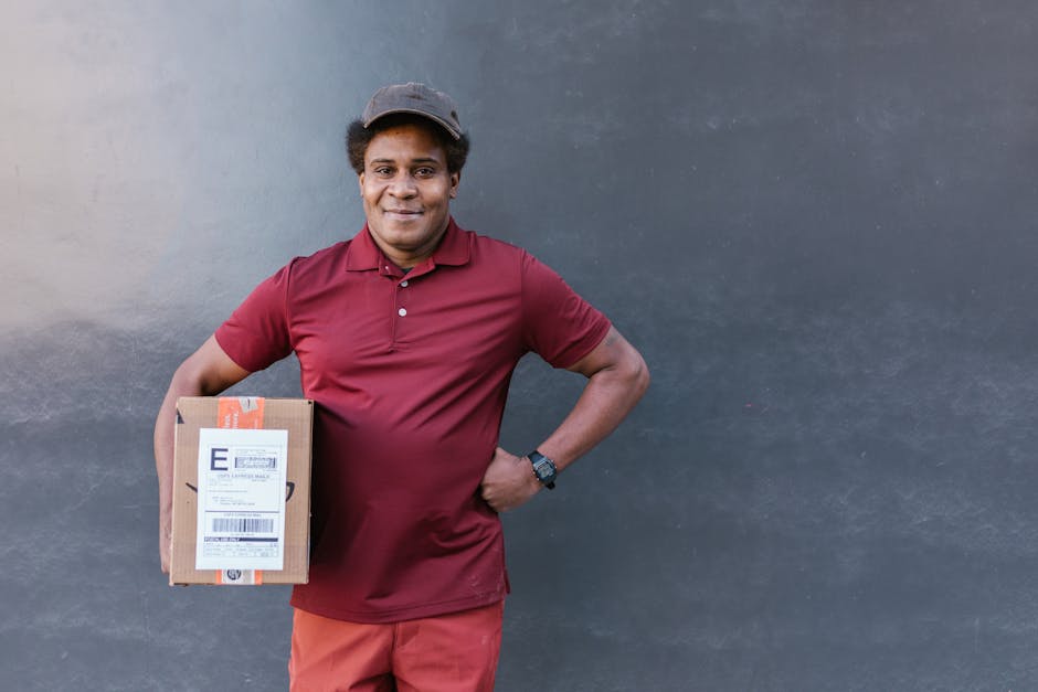 A smiling delivery person in a maroon polo shirt, orange trousers, and a dark baseball cap stands against a plain, dark grey wall. He holds a brown cardboard parcel with a white shipping label in his 