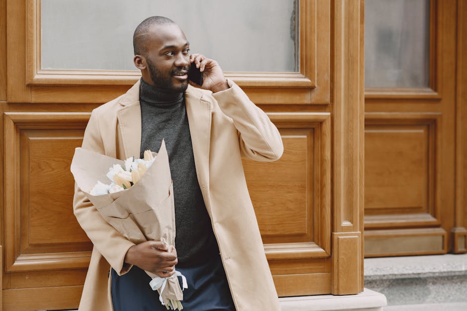 A man with dark skin and a trimmed beard stands outdoors, holding a bouquet of fresh flowers wrapped in light brown paper. He is wearing a beige overcoat over a dark grey turtleneck, with casual trous