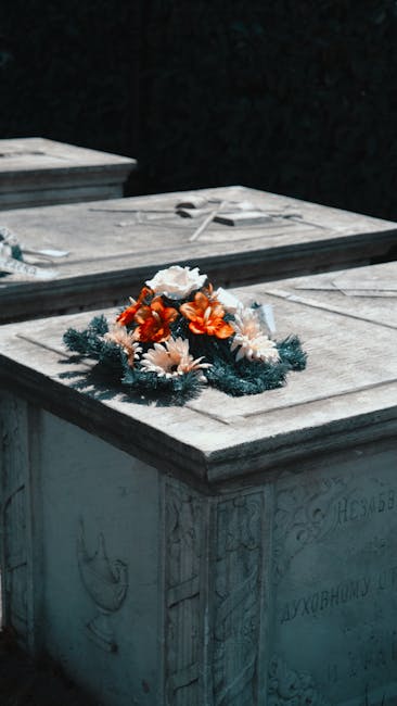 A floral tribute featuring a mixed bouquet arrangement placed atop a stone grave.The bouquet includes orange and white flowers such as daisies and roses, accented with green pine-like foliage. It is w