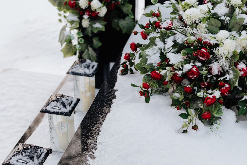 A floral arrangement featuring red roses and white flowers, partially covered in snow, resting on a table near three white candles in glass holders. The flowers are lush with green leaves, and the arr