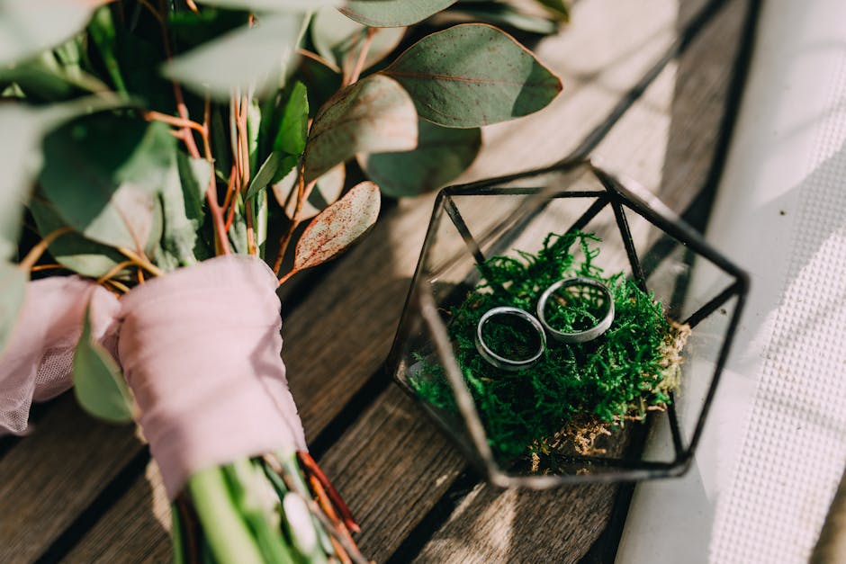 A close-up of a bouquet of fresh flowers resting on a wooden surface, featuring eucalyptus leaves with rounded, matte green and reddish-tinged foliage tied together with soft pink wrapping paper. Besi